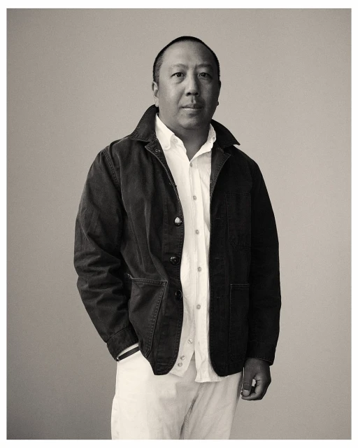 Portrait of an adult man with short dark hair, wearing a white shirt, dark jacket, and light-colored trousers, against a neutral background.