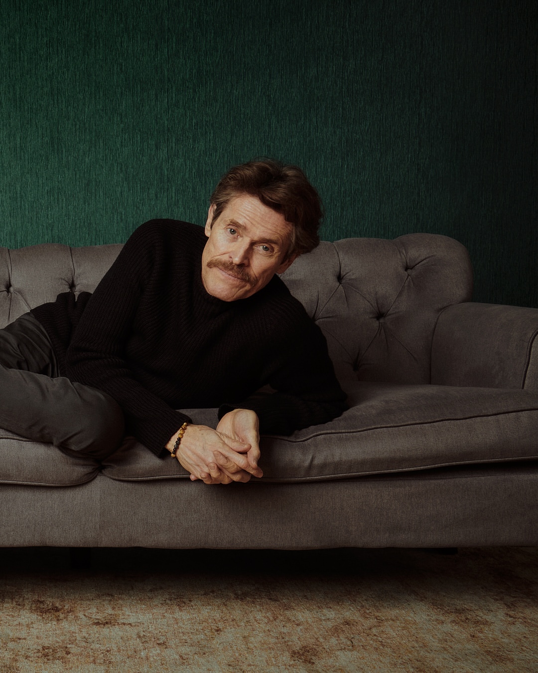 Willem Dafoe, an adult male with a mustache, rests on a tufted gray couch in a relaxed pose in a studio portrait with a dark green textured wall.