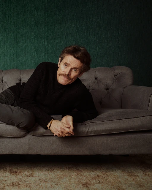 Willem Dafoe, an adult male with a mustache, rests on a tufted gray couch in a relaxed pose in a studio portrait with a dark green textured wall.