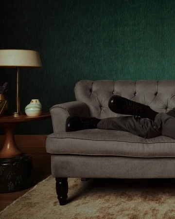 Person relaxing on a tufted grey sofa with feet up, beside a small side table and lamp against a green textured wall.