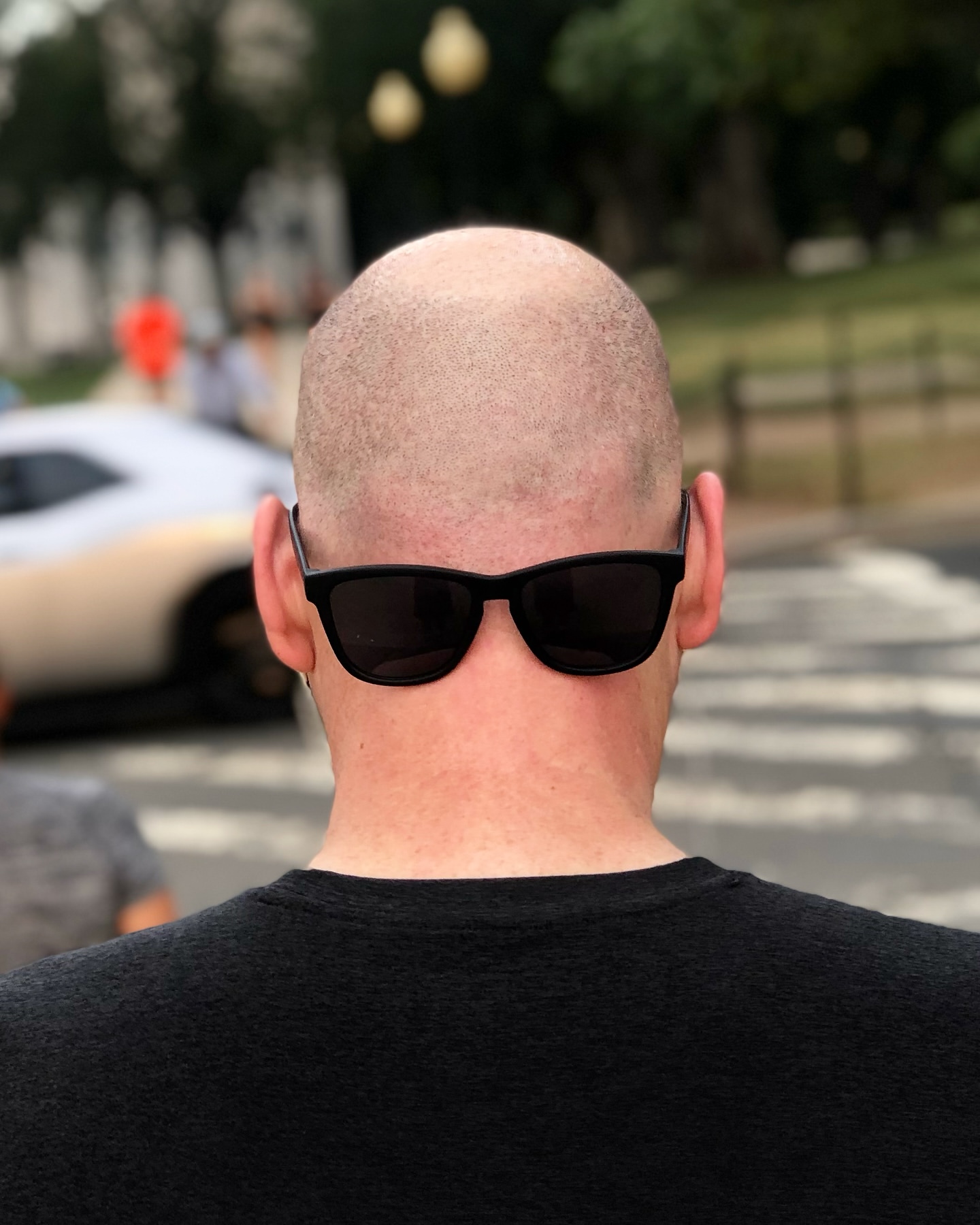 From behind, a bald man with sunglasses is walking down a crosswalk outdoors, with buildings and blurred cars in the background.