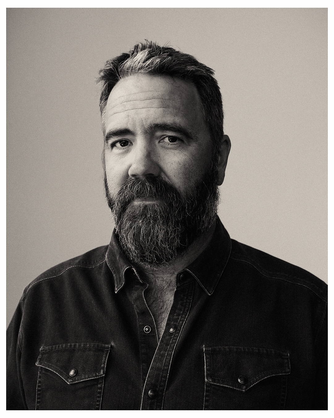 Monochrome portrait of a bearded adult male in a dark denim shirt against a neutral studio backdrop, captured with soft, diffused lighting.