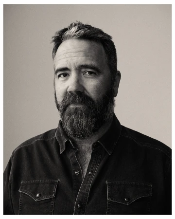 Monochrome portrait of a bearded adult male in a dark denim shirt against a neutral studio backdrop, captured with soft, diffused lighting.