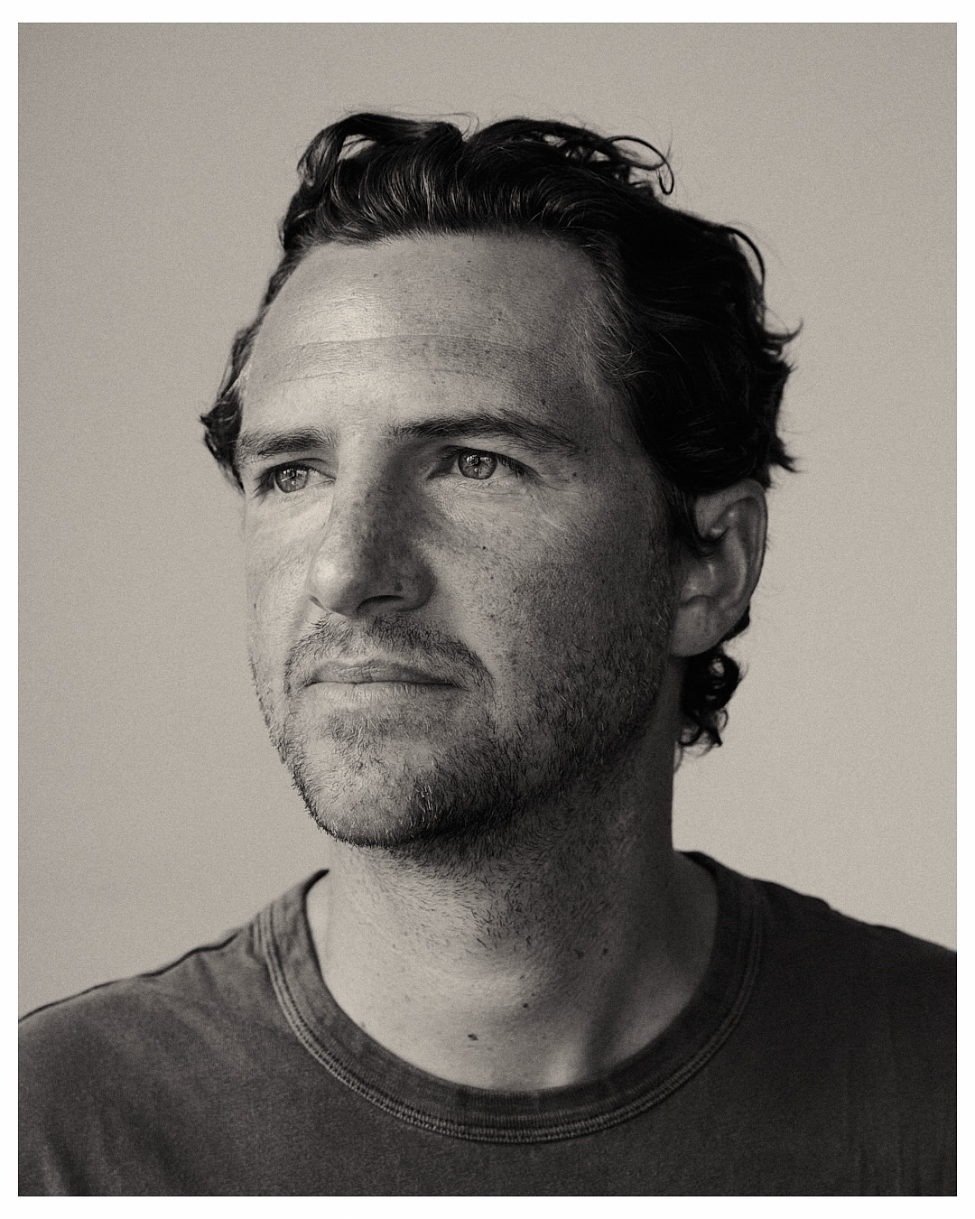 Monochrome portrait of a man with dark curly hair looking off to the side, wearing a dark t-shirt, captured in a photo studio setting.