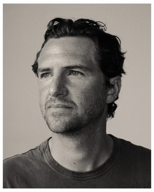 Monochrome portrait of a man with dark curly hair looking off to the side, wearing a dark t-shirt, captured in a photo studio setting.