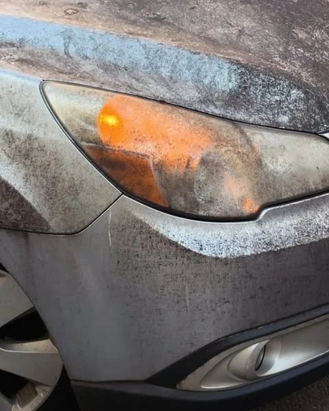 Close-up shot of a dirty silver car's headlight with the blinker on. The car's body is covered in grime, suggesting it has not been cleaned.