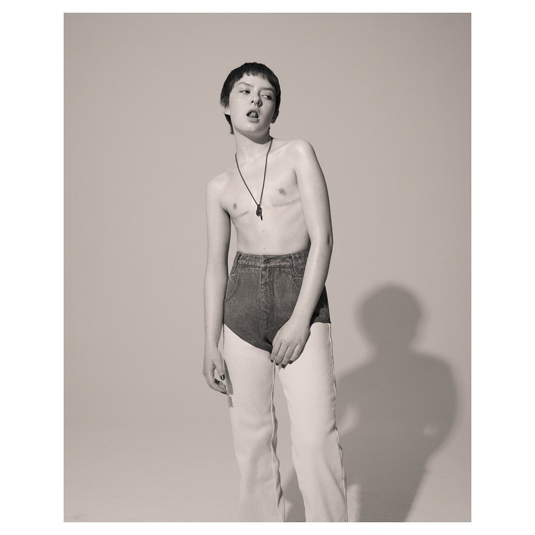 Monochrome portrait of a young adult posing shirtless with short hair, necklace, blue denim shorts and baggy light-colored jeans.