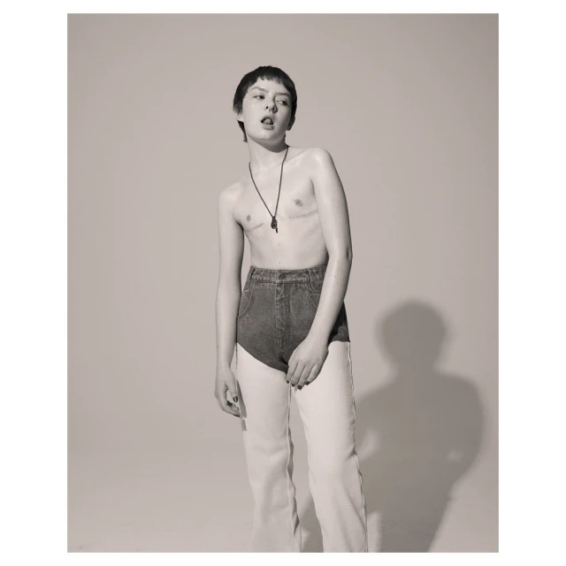 Monochrome portrait of a young adult posing shirtless with short hair, necklace, blue denim shorts and baggy light-colored jeans.