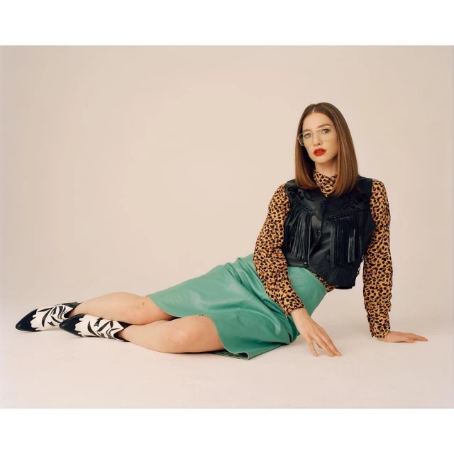 A young woman with long straight brown hair is lying on her side wearing a black fringed vest, animal print top, green skirt, and cowboy boots.