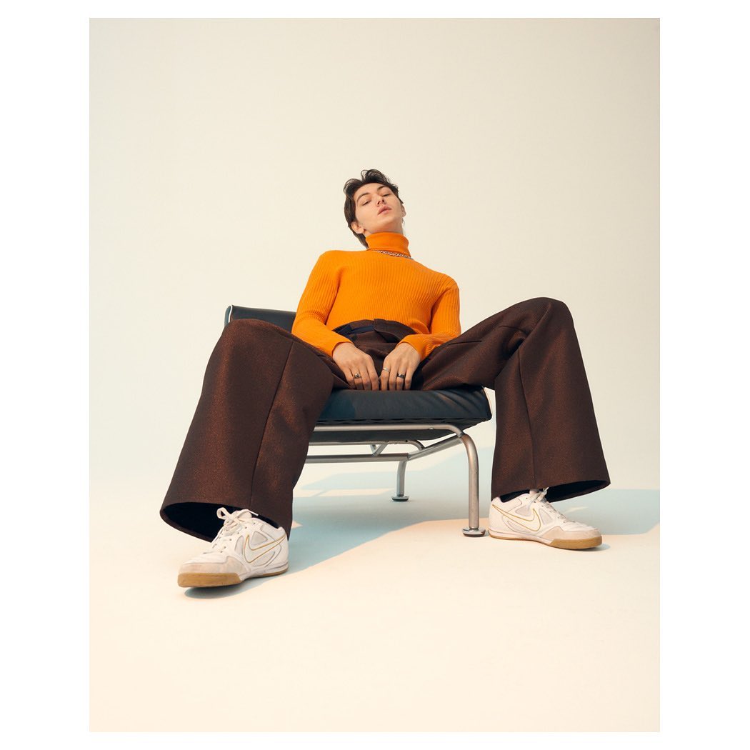 An adult male seated in a black chair, wearing an orange turtleneck sweater, brown pants, and white sneakers against a light background.