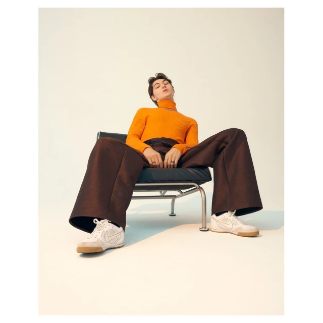 An adult male seated in a black chair, wearing an orange turtleneck sweater, brown pants, and white sneakers against a light background.