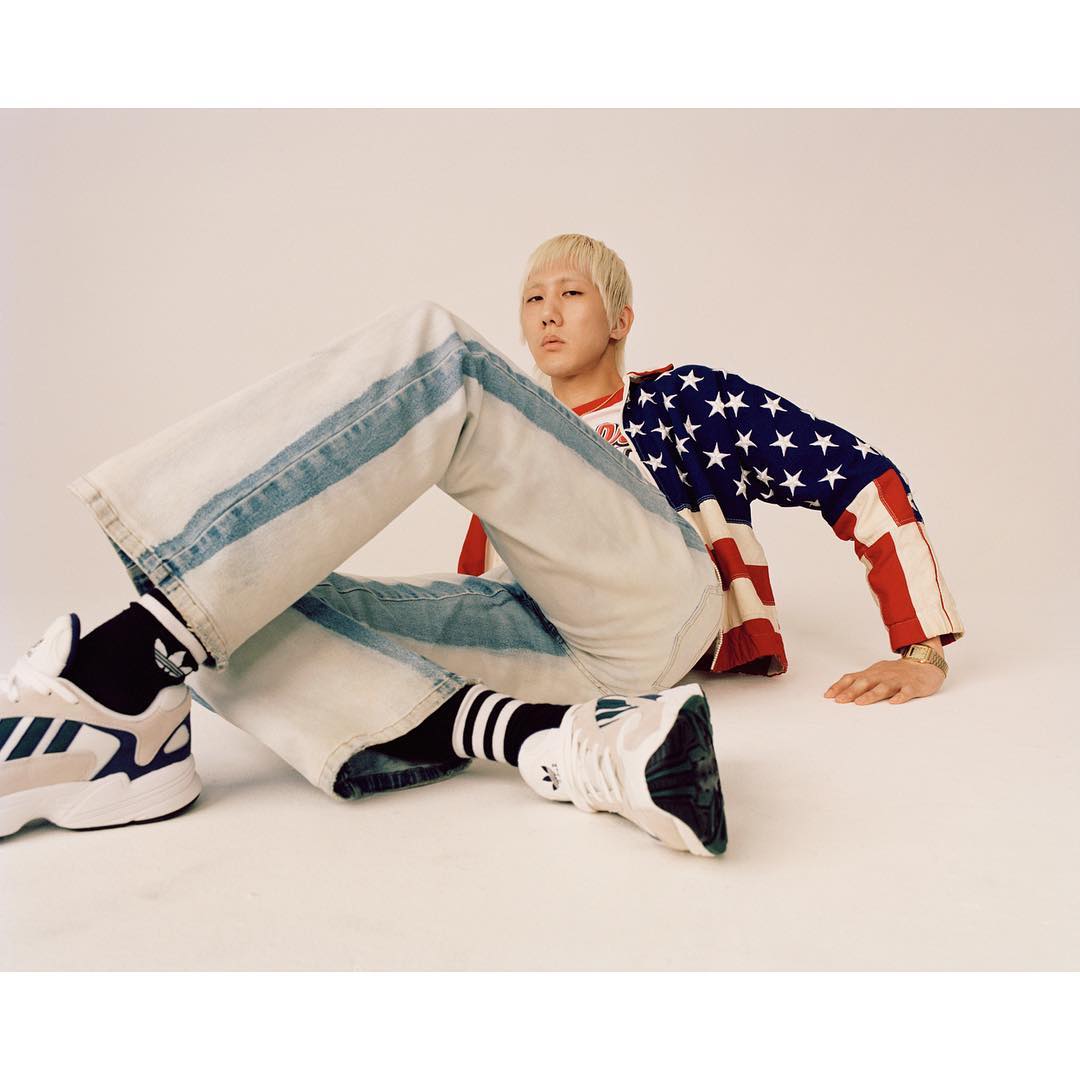 Blonde man wearing an American flag shirt and jeans poses with Adidas athletic shoes in a photo studio on a neutral background.
