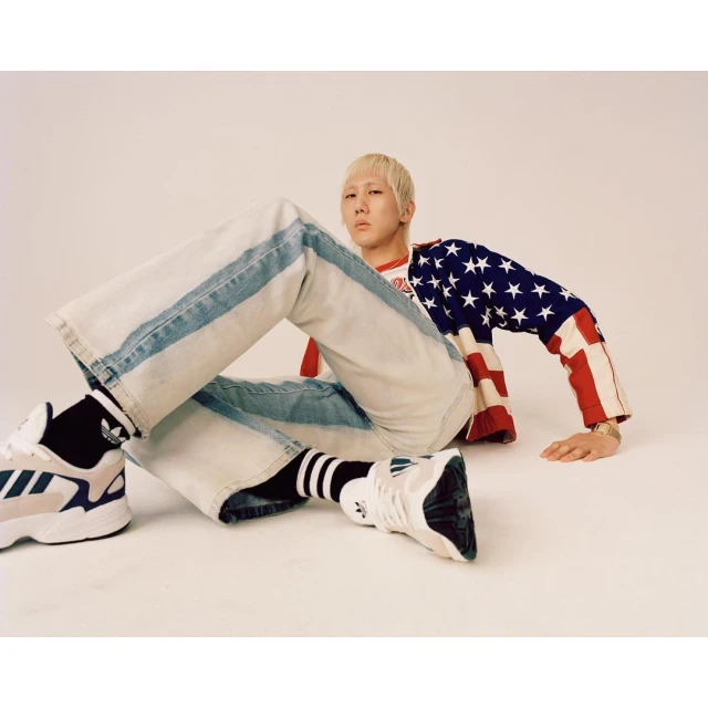 Blonde man wearing an American flag shirt and jeans poses with Adidas athletic shoes in a photo studio on a neutral background.