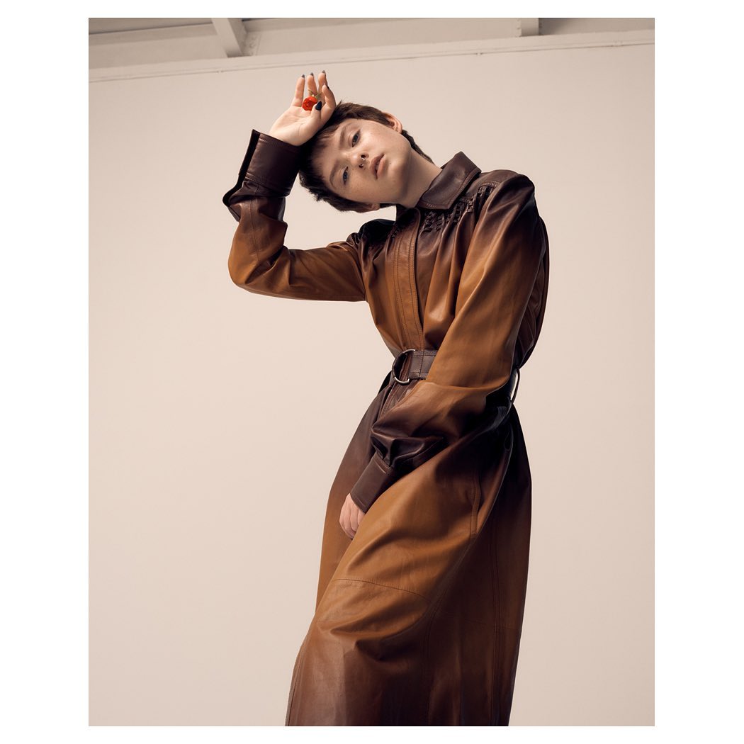 Woman in a brown leather coat poses indoors with a tilted head and raised hand, creating an editorial fashion shot against a plain backdrop.