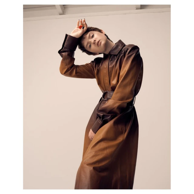 Woman in a brown leather coat poses indoors with a tilted head and raised hand, creating an editorial fashion shot against a plain backdrop.