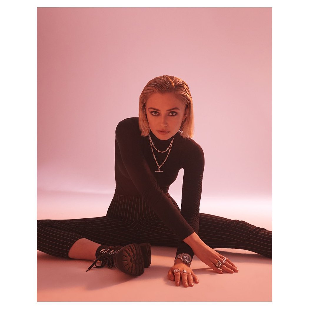 Blonde woman poses sitting on the floor in black turtleneck, pinstripe pants, platform boots, jewelry and watch in pink-toned photo studio.