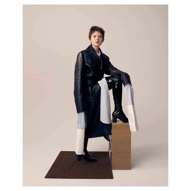 Full-length fashion shot of a young adult woman in a black leather coat and boots, posing on a wooden crate in a photo studio.