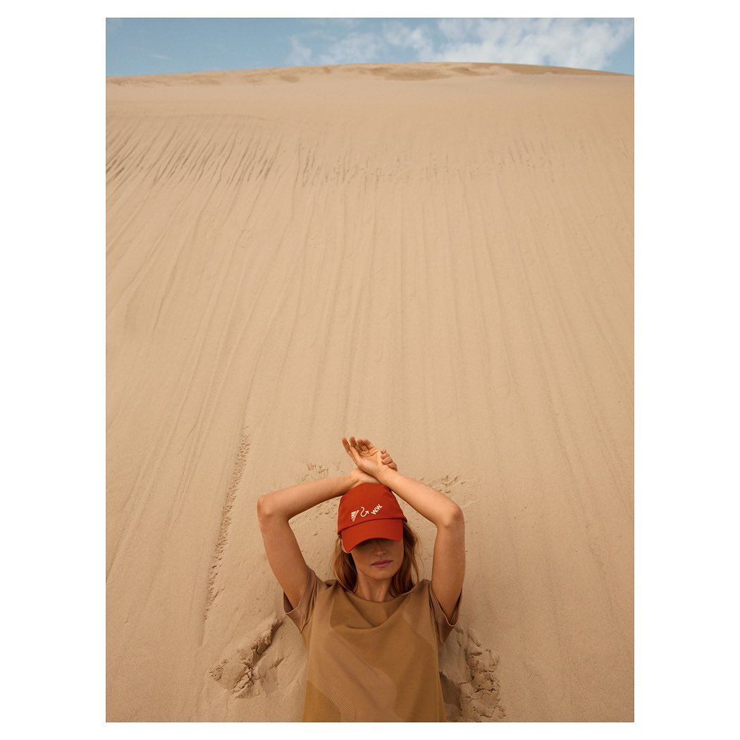Woman in red cap lies in sand with arms overhead. Dune backdrop. Neutral color palette, fashion-focused.