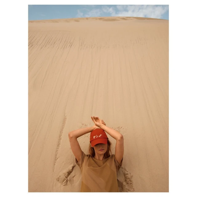 Woman in red cap lies in sand with arms overhead. Dune backdrop. Neutral color palette, fashion-focused.