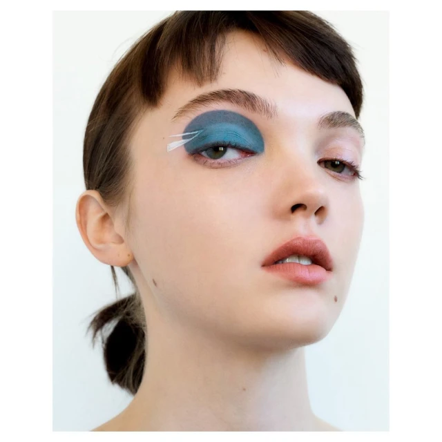 Close-up portrait of a young adult woman wearing bold blue eyeshadow and white feather accents for a creative beauty look.