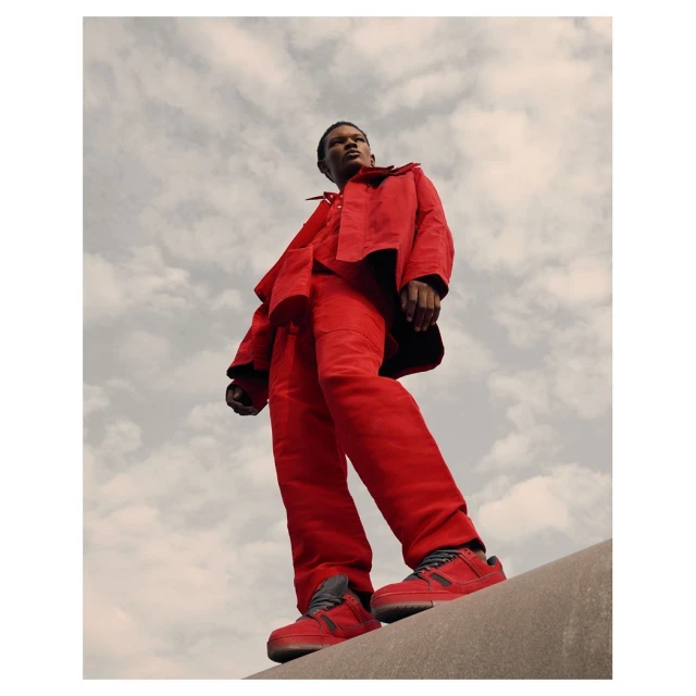 A striking low-angle shot of a Black male model in red outerwear and pants, standing on a concrete ledge against a cloudy sky.