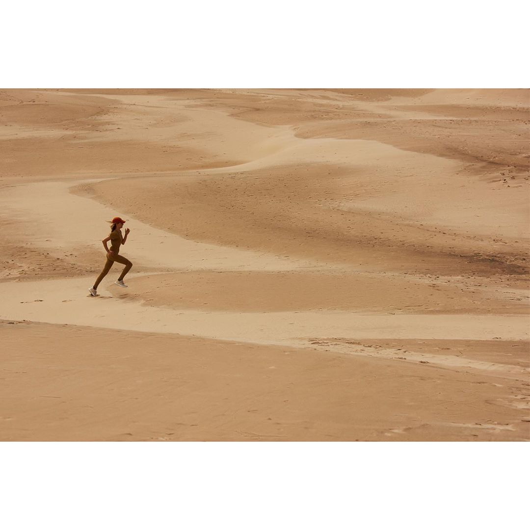 A woman is running across a desert landscape with sand dunes, wearing a red hat, tan athletic suit, and sneakers, during the daytime.
