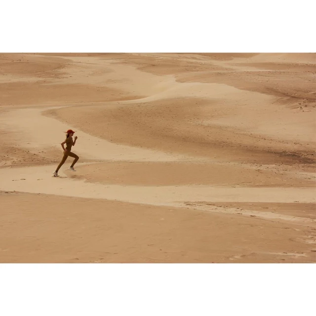 A woman is running across a desert landscape with sand dunes, wearing a red hat, tan athletic suit, and sneakers, during the daytime.