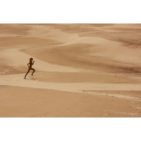 A woman is running across a desert landscape with sand dunes, wearing a red hat, tan athletic suit, and sneakers, during the daytime.