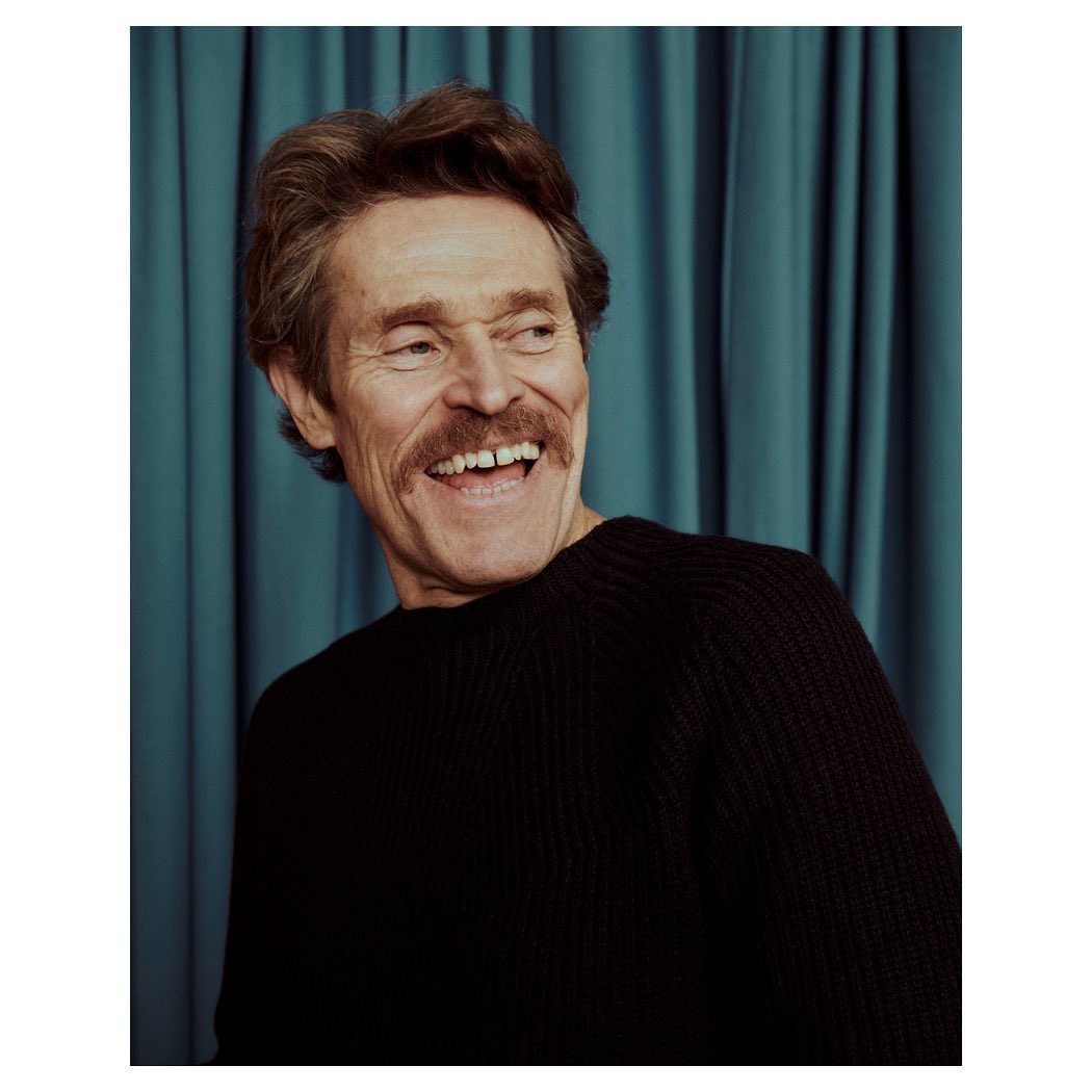 Close-up portrait of a smiling adult man with a mustache, wearing a black sweater, set against a teal curtain backdrop.