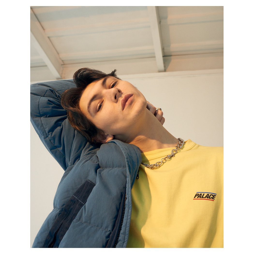 Portrait of a young adult with dark hair wearing a yellow Palace t-shirt and blue puffer jacket indoors, with a silver chain necklace.