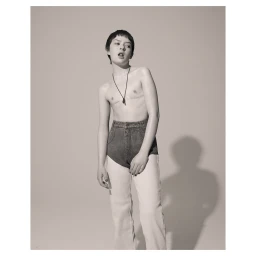 Portrait of a person with short dark hair wearing high-waisted denim pants against a plain studio background.