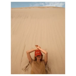 Woman wearing a red baseball cap and tan t shirt posing against a vast sandy desert dune under a clear sky