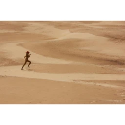 A lone figure running across vast undulating sand dunes under bright natural light.