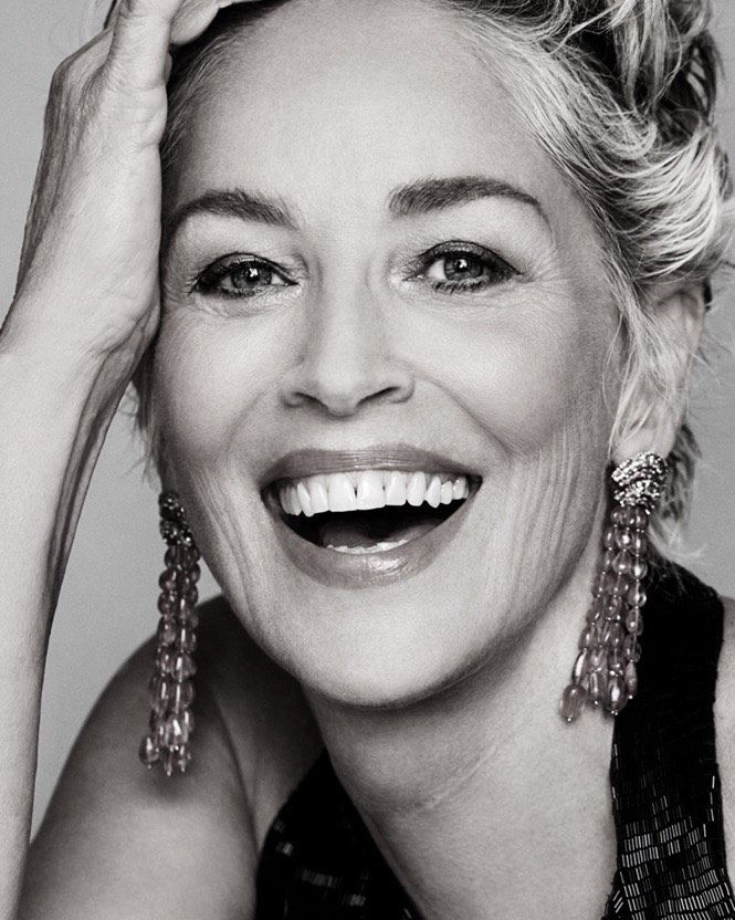 Close-up monochrome portrait of Sharon Stone smiling broadly with hand to her head, wearing beaded earrings and a dark sleeveless top.