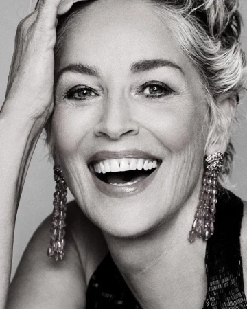 Close-up monochrome portrait of Sharon Stone smiling broadly with hand to her head, wearing beaded earrings and a dark sleeveless top.
