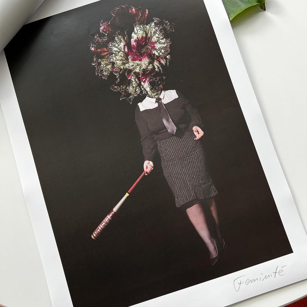 Surreal portrait of person with flower head in dark clothing holding a baseball bat in a studio setting against a black background.