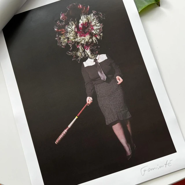 Surreal portrait of person with flower head in dark clothing holding a baseball bat in a studio setting against a black background.