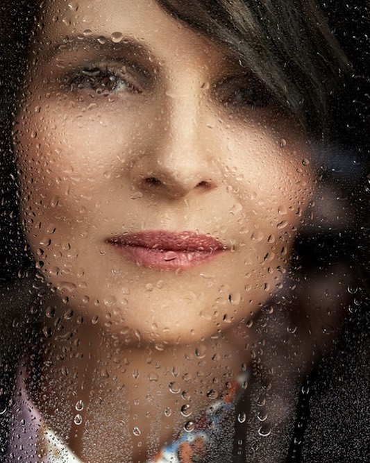 Close-up portrait of a middle-aged woman with dark hair behind a window covered in water droplets, with a blurred background.