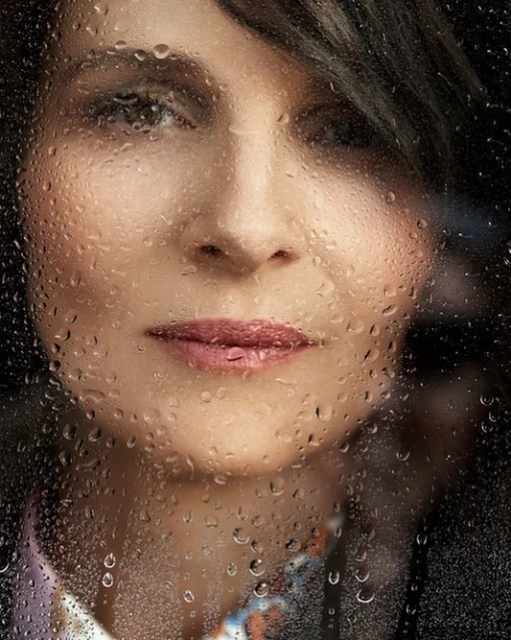 Close-up portrait of a middle-aged woman with dark hair behind a window covered in water droplets, with a blurred background.
