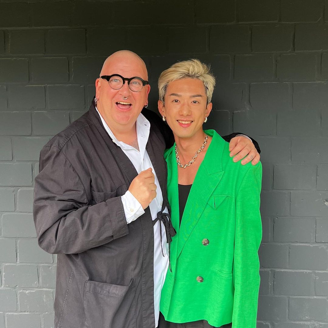Two adult men pose against a gray brick wall. One man wears a black robe and glasses, while the other sports a green blazer and silver chain.