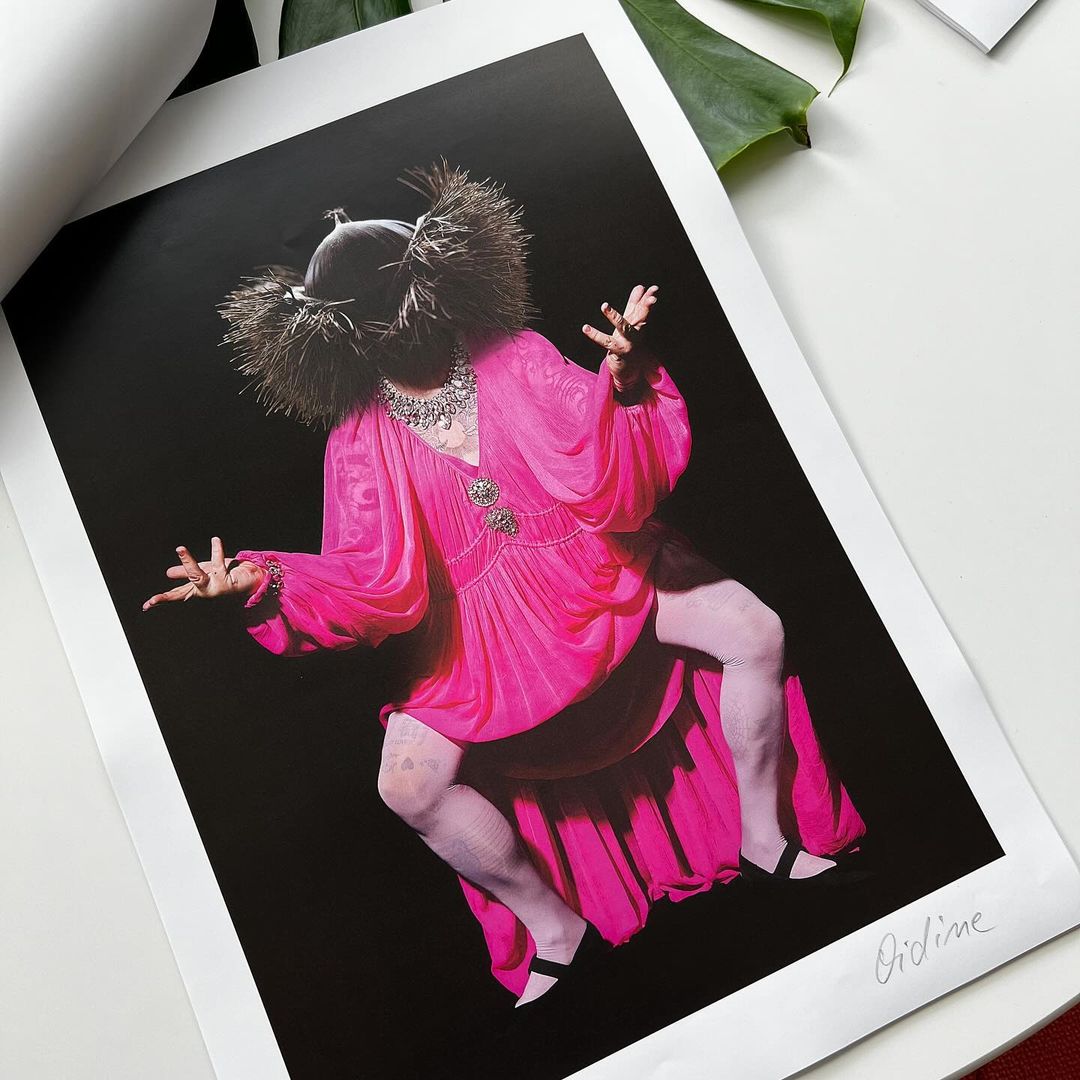 Artwork featuring a person in a bright pink dress with decorative headwear against a black backdrop, presented as a framed print on a white table.