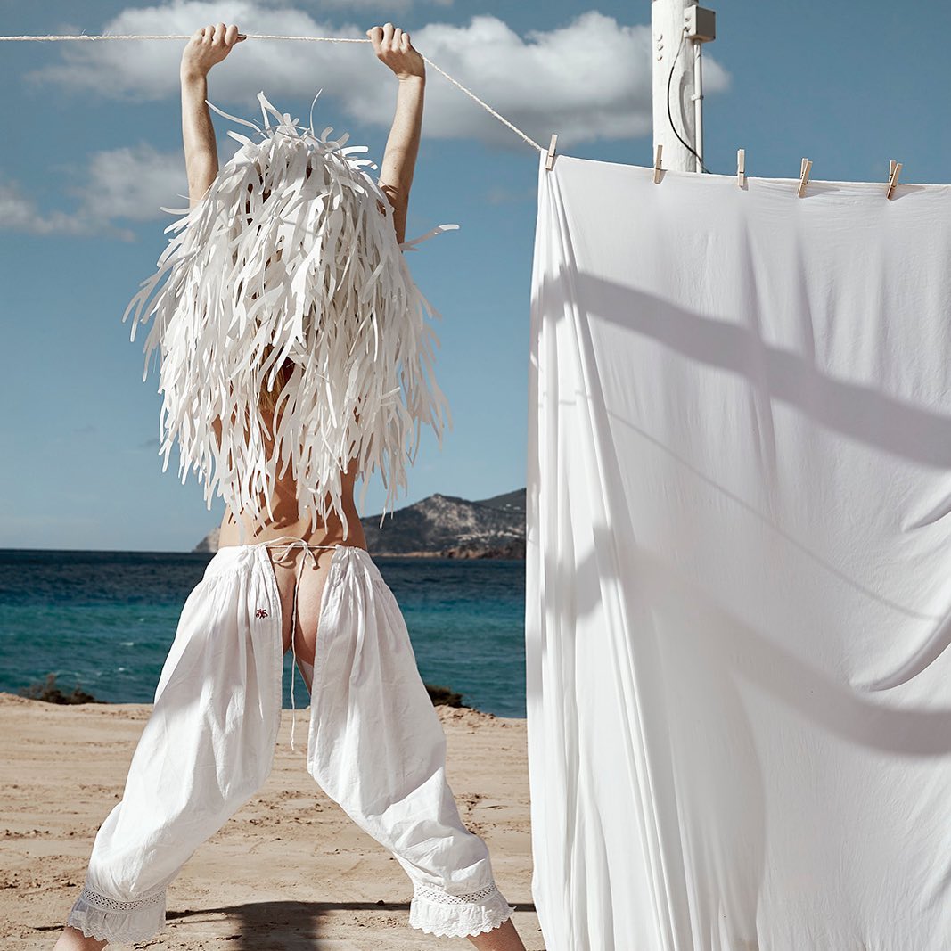 Woman in baggy white pants and decorative headpiece, stands on a sandy beach with turquoise water in the background, holding a clothesline.