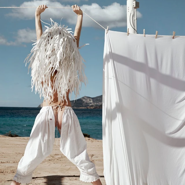 Woman in baggy white pants and decorative headpiece, stands on a sandy beach with turquoise water in the background, holding a clothesline.