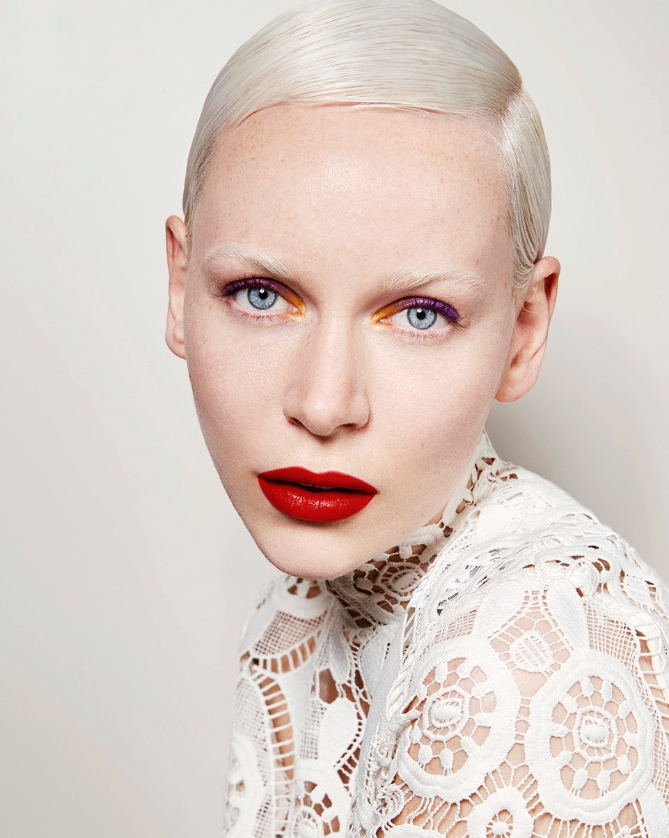 Close-up studio shot of a female model with platinum blonde hair, bright blue eyes, red lips, and unique purple and gold eye makeup.