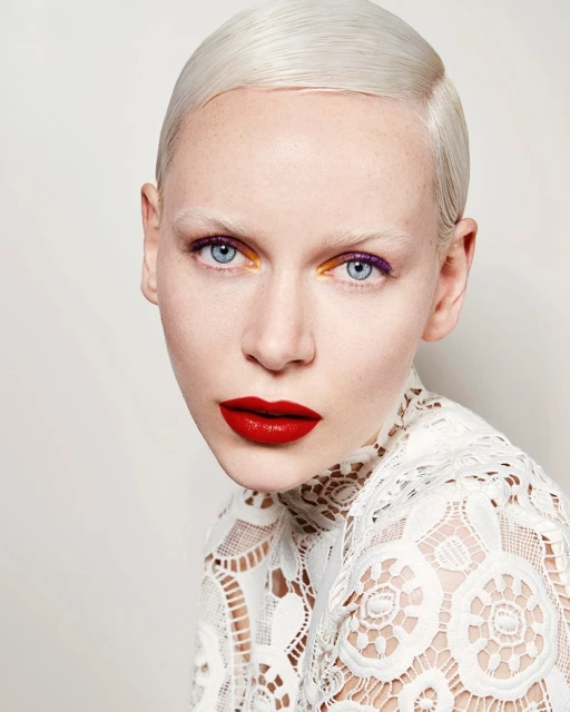 Close-up studio shot of a female model with platinum blonde hair, bright blue eyes, red lips, and unique purple and gold eye makeup.