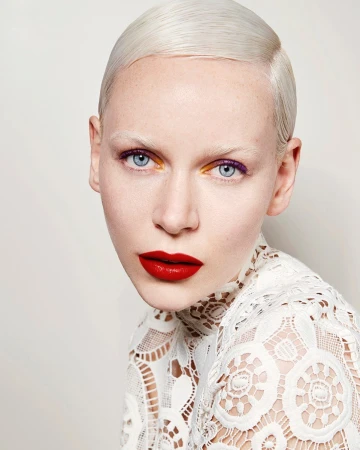 Close-up studio shot of a female model with platinum blonde hair, bright blue eyes, red lips, and unique purple and gold eye makeup.