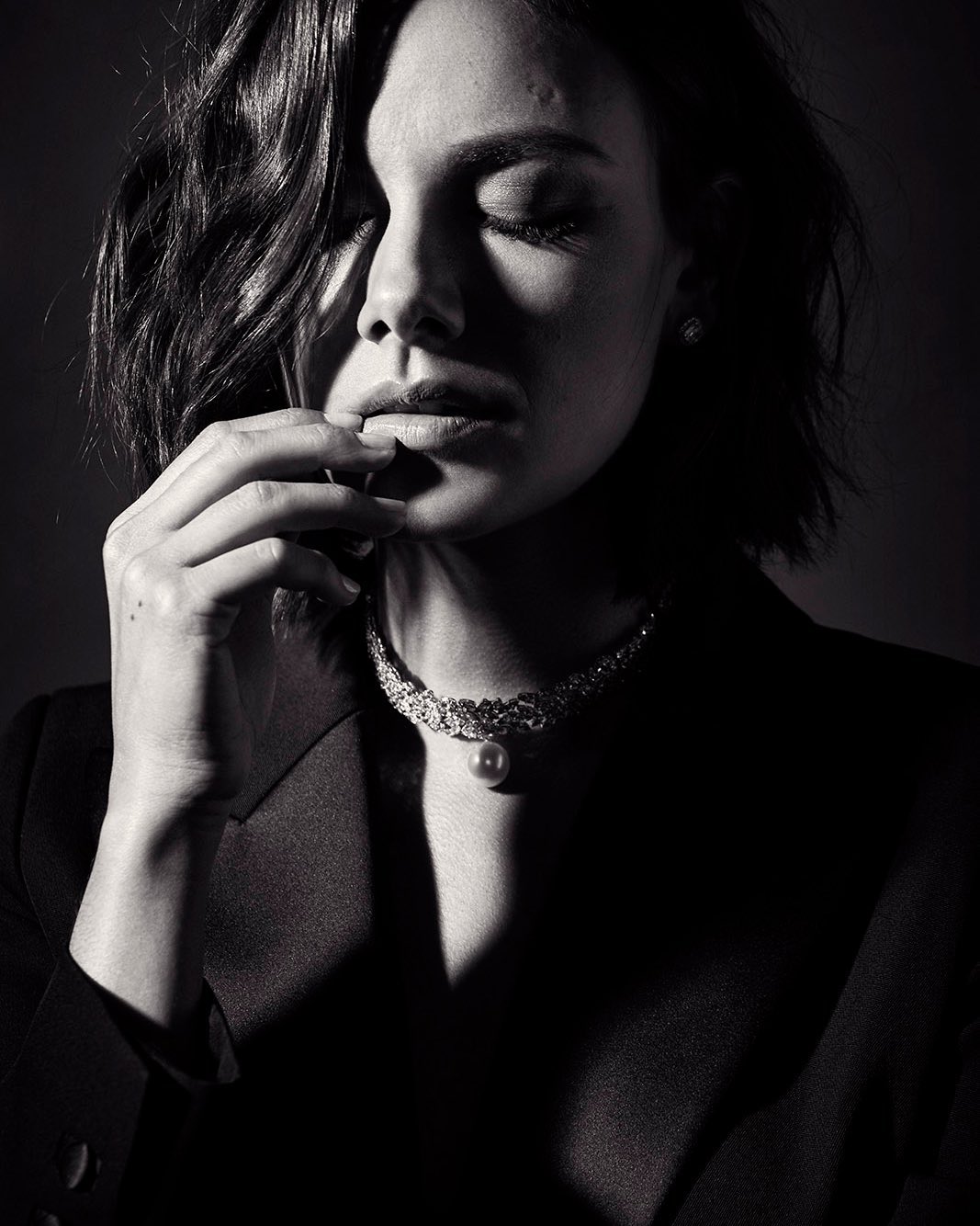 Monochrome portrait of a woman with her eyes closed, wearing a dark blazer and an elegant necklace, hand touching her lips.