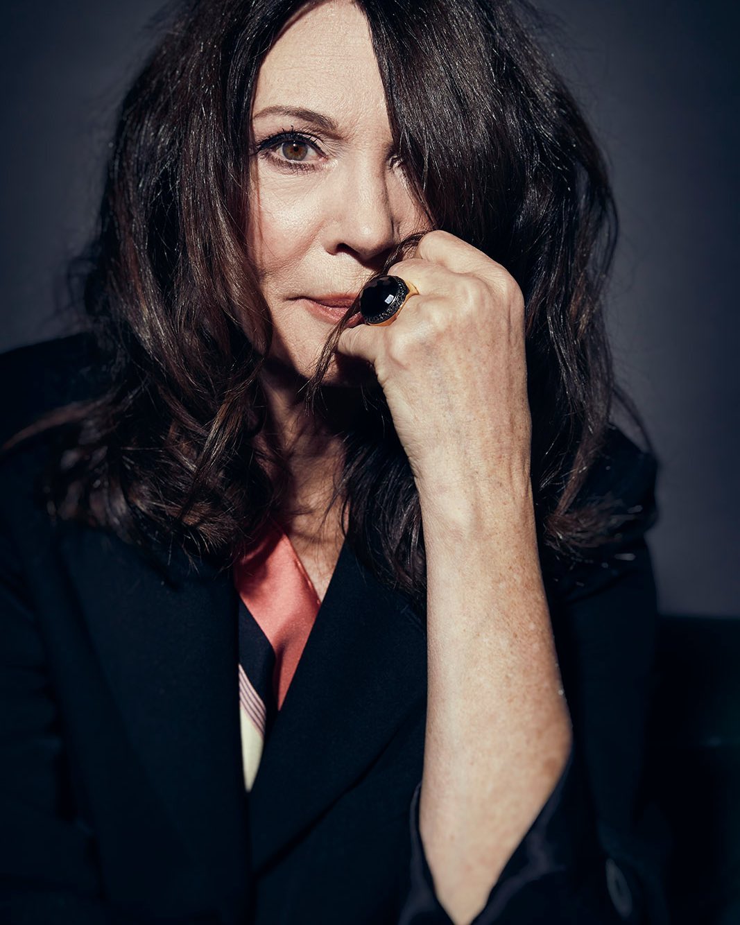 Portrait of a brunette woman wearing a black blazer and an oversized ring, posing in front of a dark background.