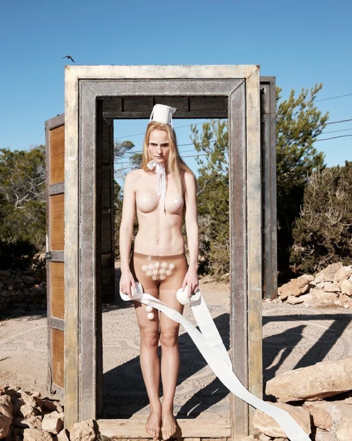 A nude woman stands in a doorway outdoors, wearing a makeshift hat and holding a roll of toilet paper, creating an odd and intriguing scene.