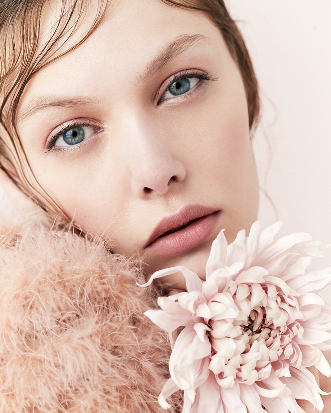 Close-up portrait of a fair-skinned young woman with blue eyes and wet-look hair holding a large pink dahlia flower against a pale pink background.